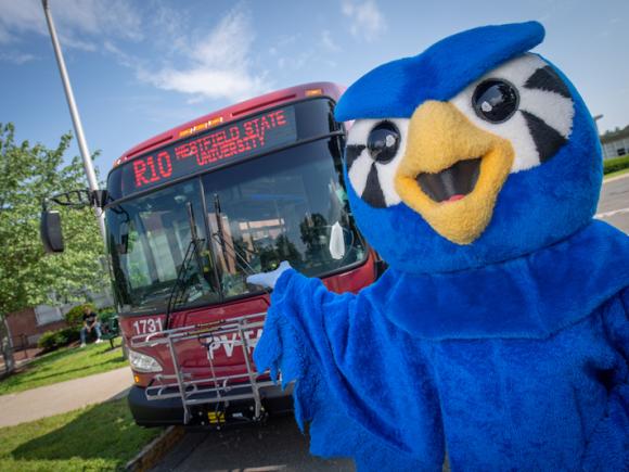 A photo of Nestor in his blue, owl costume and pointing towards a red bus which has the university's address and route on the front.