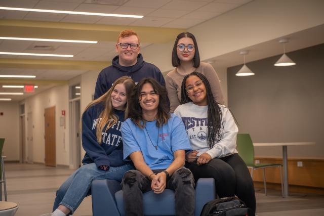 Five students wearing 蓝莓视频 State sweatshirts smiling in Parenzo Hall. Two standing three sitting.