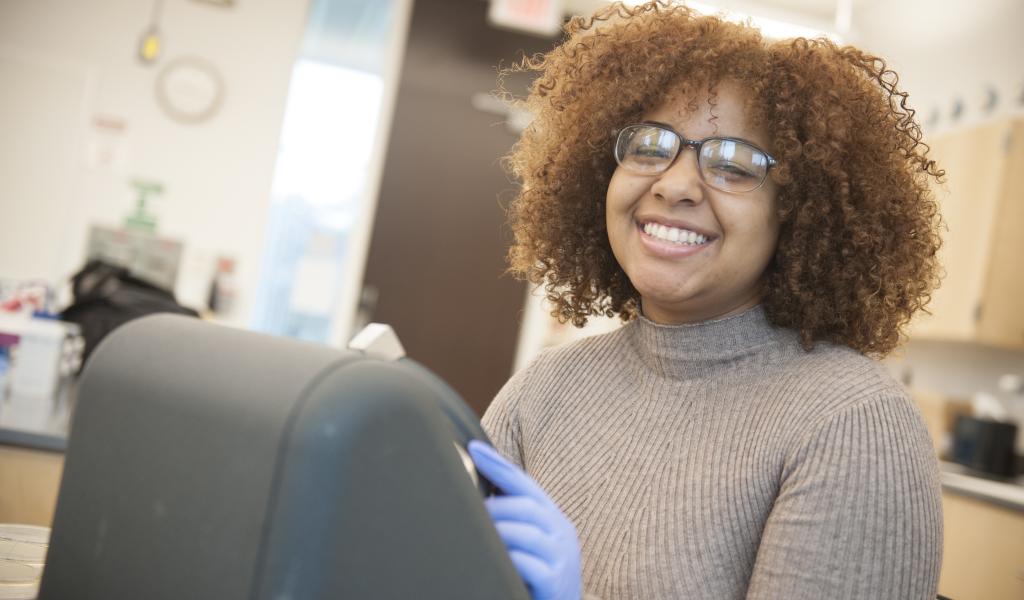 female student with curly hair and glasses smiles at the camera as she prepares samples in the lab